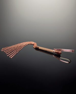 A flogger with a brown handle and numerous copper-colored chains, resting on a reflective dark surface.