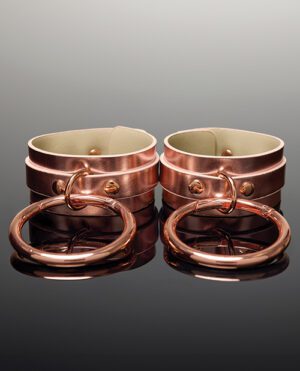 A pair of polished copper-coloured cuffs with large circular rings attached, displayed on a reflective surface with a gradient background.