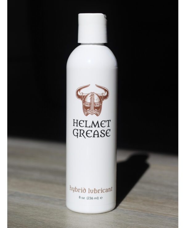 A white bottle labeled 'HELMET GREASE hybrío lubricant' with a viking helmet logo, standing against a black background with a shadow cast on a wooden surface.