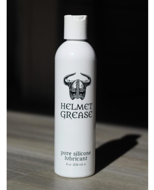 A white bottle of pure silicone lubricant labeled 'Helmet Grease' with a viking helmet logo, standing on a wooden surface against a dark background.