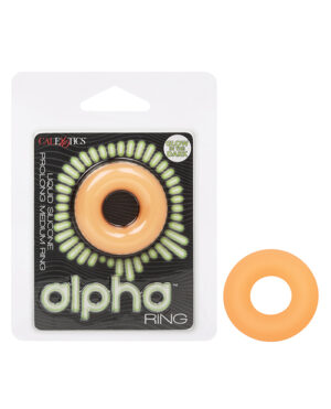 The image shows a product package for an "Alpha Ring" by CalExotics. The ring is positioned in the center with a glow-in-the-dark feature highlighted on the package. The packaging is white with black and green text and accents. A loose ring, matching the one inside the packaging, is displayed to the right, showcasing its orange color and circular shape.