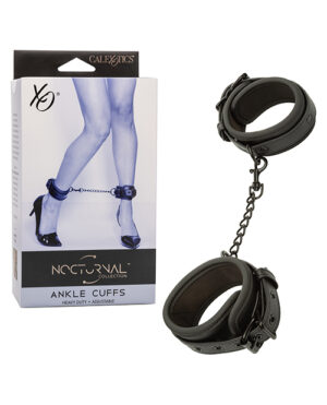 A pair of black ankle cuffs with a connecting chain next to their product box which features an image of a pair of legs wearing the cuffs. The box is labeled "Nocturnal Collection Ankle Cuffs" by CalExotics.