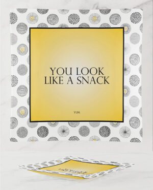 A decorative piece of wall art with the phrase "YOU LOOK LIKE A SNACK" in a yellow square frame, with a patterned background and a matching booklet on a marble surface.