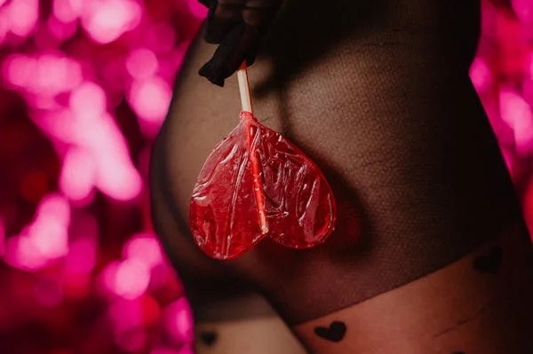 A close-up image of a person holding a red heart-shaped lollipop while wearing black stockings with heart patterns, set against a vibrant pink backdrop.